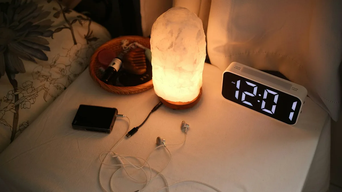 Despertador digital ao lado de um abajur em uma mesa de cabeceira, objeto comum que também existe como câmera espiã
