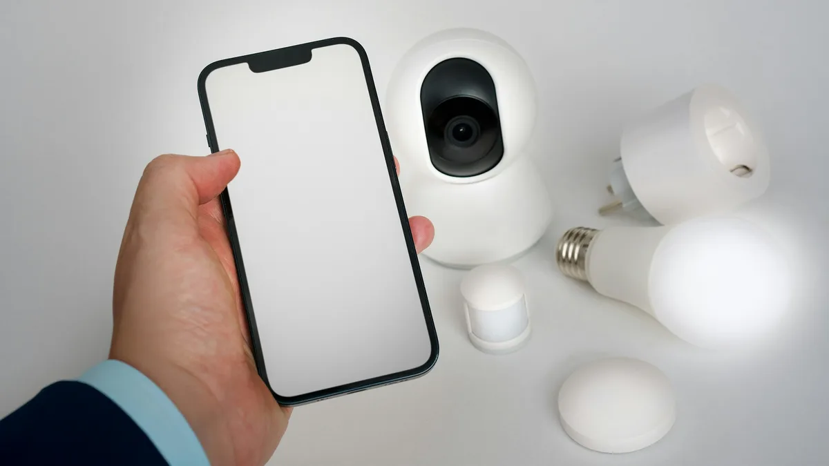 Mão segurando celular ao lado de câmera Wi-Fi branca e outros dispositivos de casa conectada