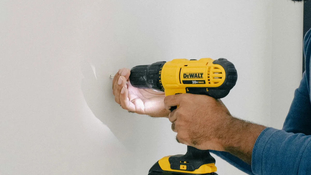 Homem furando uma parede branca com furadeira amarela para instalar suporte