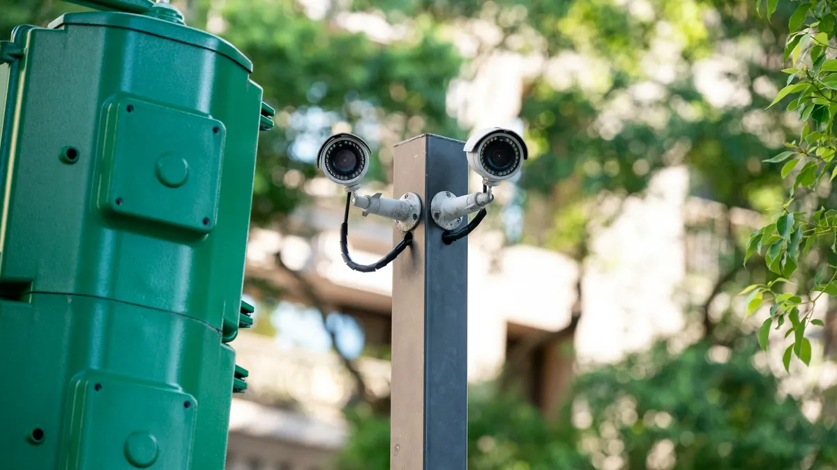 Duas câmeras de segurança presas a um poste ao lado de uma caixa de passagem verde, no meio das árvores